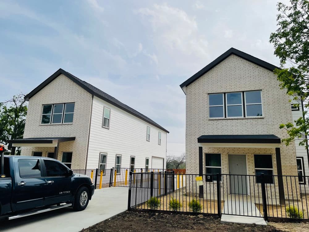 Two modern houses with a truck parked in front, featuring a fenced yard and clear skies.